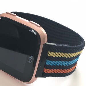 May include: A rose gold Fitbit smartwatch with a black and multi-colored striped elastic band. The band has yellow, blue, and orange stripes.
