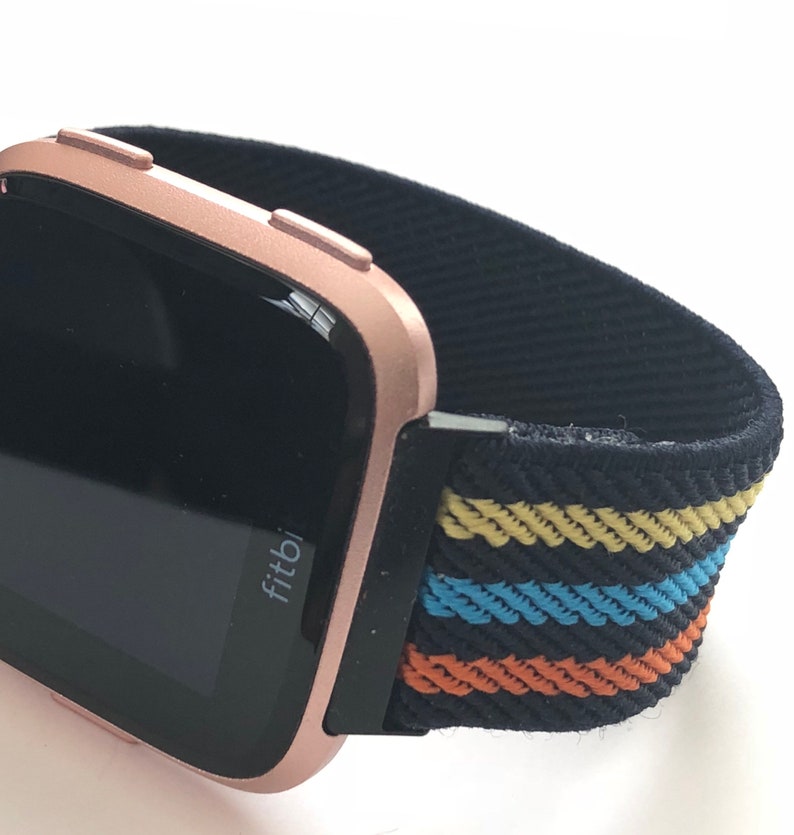 May include: A rose gold Fitbit smartwatch with a black and multi-colored striped elastic band. The band has yellow, blue, and orange stripes.