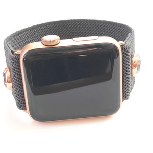 May include: A rose gold Apple Watch with a black sport band. The band has a textured fabric design and features small rhinestones on each side.