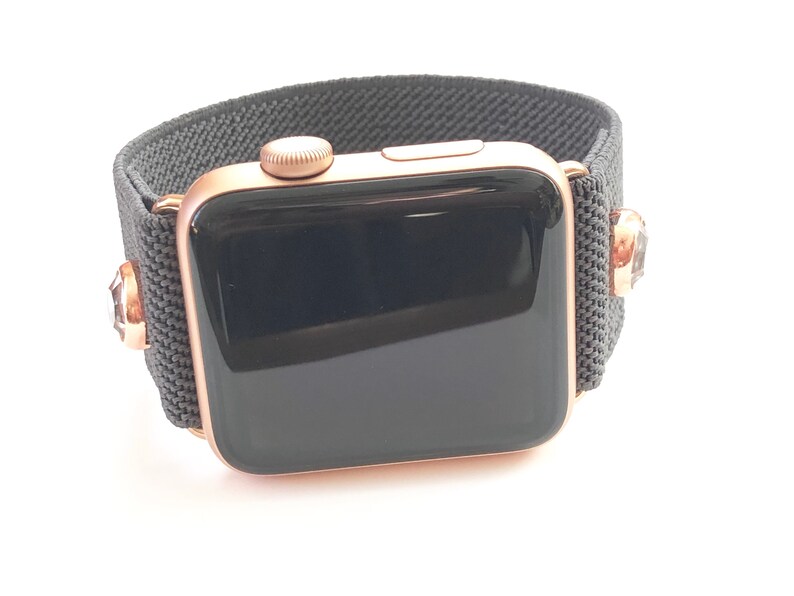 May include: A rose gold Apple Watch with a black sport band. The band has a textured fabric design and features small rhinestones on each side.
