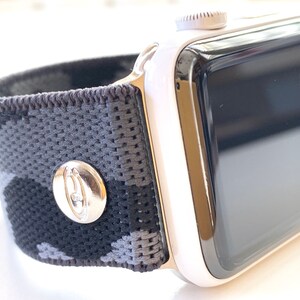 May include: A white Apple Watch with a black and grey camouflage patterned sport band. The band has a silver buckle with a logo.