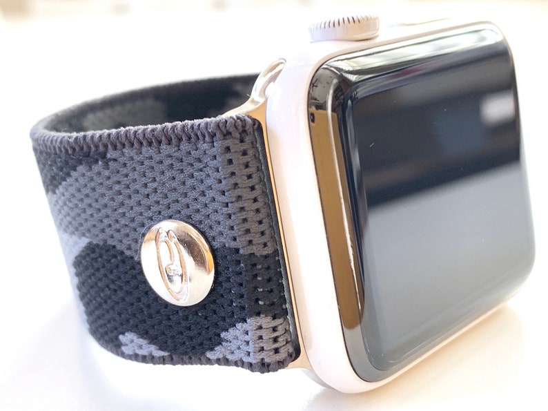 May include: A white Apple Watch with a black and grey camouflage patterned sport band. The band has a silver buckle with a logo.