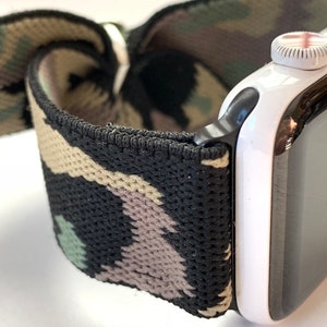 May include: A black and green camouflage patterned elastic watch band attached to a white Apple Watch with a black bezel.