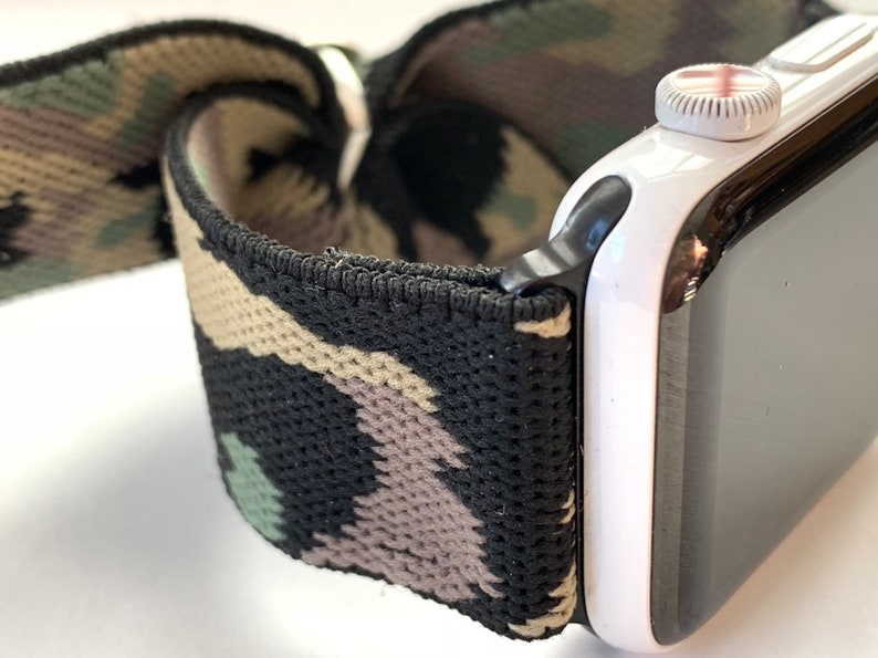May include: A black and green camouflage patterned elastic watch band attached to a white Apple Watch with a black bezel.