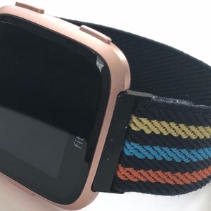 May include: Rose gold Fitbit smartwatch with a black and multi-colored striped fabric band. The watch face displays the word "Fit".