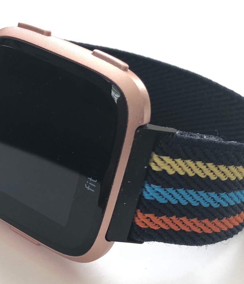 May include: Rose gold Fitbit smartwatch with a black and multi-colored striped fabric band. The watch face displays the word "Fit".