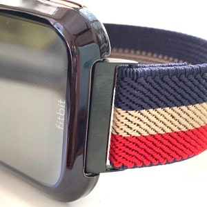 Puede incluir: Un reloj inteligente Fitbit negro con una banda el&aacute;stica a rayas azul marino, beige y rojo. La banda tiene un cierre de metal negro.