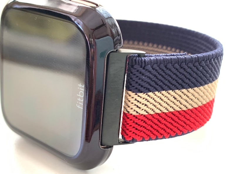 Puede incluir: Un reloj inteligente Fitbit negro con una banda el&aacute;stica a rayas azul marino, beige y rojo. La banda tiene un cierre de metal negro.