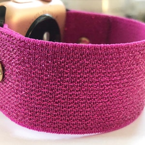 May include: A pink glitter watch band with a clear rhinestone accent. The band is made of a stretchy material and is designed to fit a smartwatch.