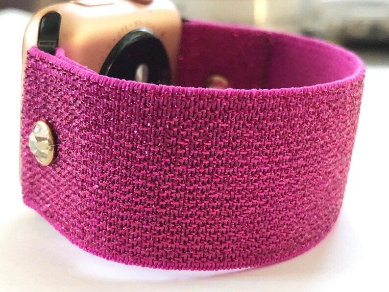 May include: A pink glitter watch band with a clear rhinestone accent. The band is made of a stretchy material and is designed to fit a smartwatch.