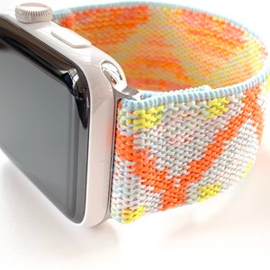 Pode incluir: Um Apple Watch branco com uma face preta e uma banda tecida colorida. A banda é uma mistura de laranja, amarelo e azul.