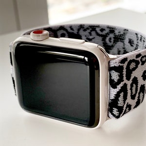 Puede incluir: Un Apple Watch blanco con una banda deportiva de estampado de leopardo blanco y negro. La esfera del reloj es negra y la corona digital es roja.