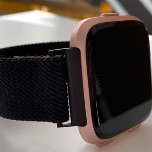 May include: A rose gold Fitbit smartwatch with a black woven band. The watch face is black and displays the Fitbit logo.