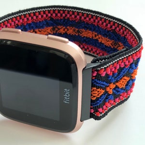 May include: A rose gold Fitbit smartwatch with a black and colorful patterned elastic band. The band has a geometric design with red, orange, blue, and white accents.