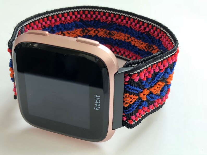 May include: A rose gold Fitbit smartwatch with a black and colorful patterned elastic band. The band has a geometric design with red, orange, blue, and white accents.