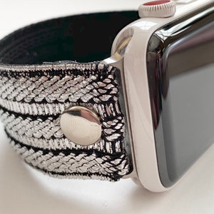 Puede incluir: Una correa de reloj plateada y negra con cierre de bot&oacute;n plateado. La correa est&aacute; hecha de tejido tejido y es compatible con Apple Watch.