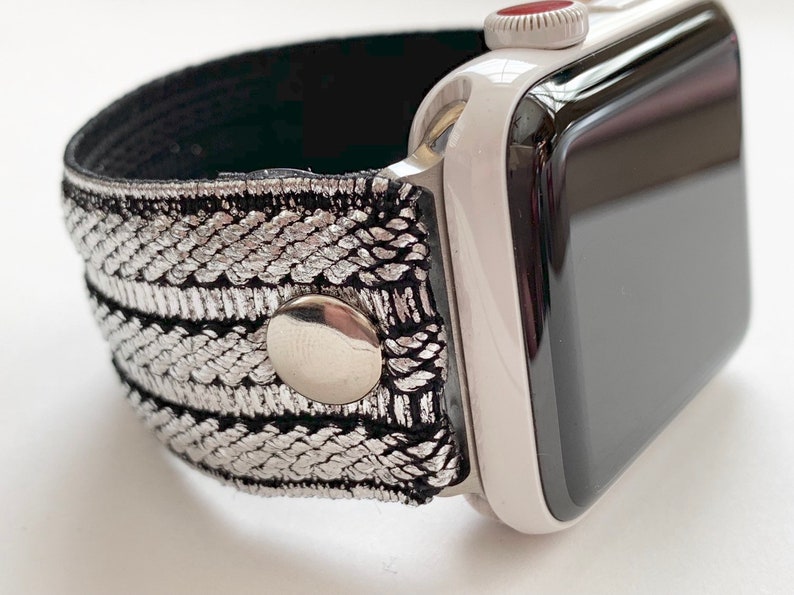 Puede incluir: Una correa de reloj plateada y negra con cierre de bot&oacute;n plateado. La correa est&aacute; hecha de tejido tejido y es compatible con Apple Watch.