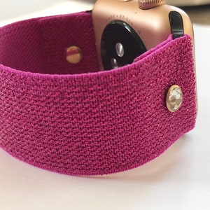 May include: A pink sparkly watch band with two gold-toned metal clasps. The band is made of a stretchy fabric and has a glittery finish.