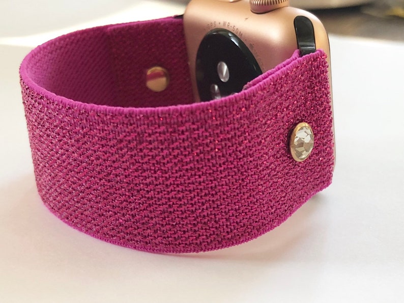 May include: A pink sparkly watch band with two gold-toned metal clasps. The band is made of a stretchy fabric and has a glittery finish.