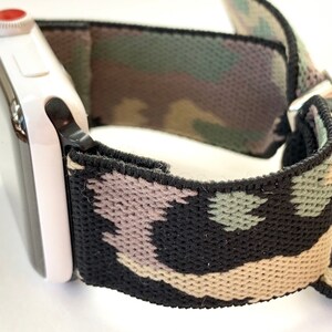 May include: A black, green, and beige camouflage patterned watch band with a white Apple Watch attached.