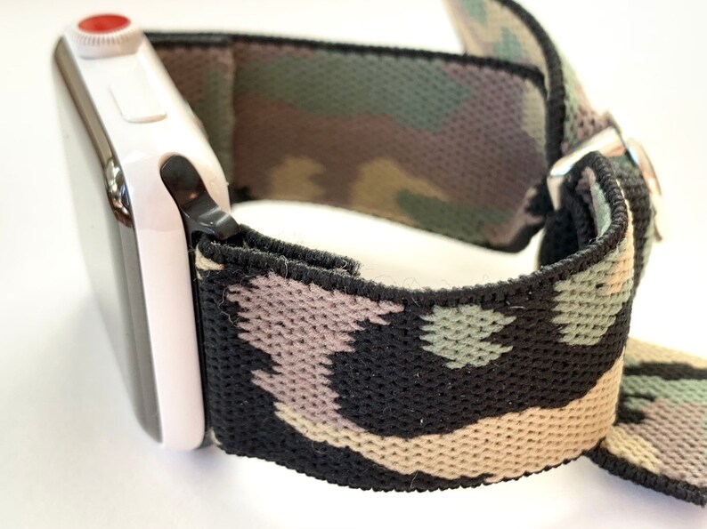 May include: A black, green, and beige camouflage patterned watch band with a white Apple Watch attached.