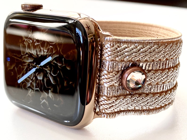 May include: A gold Apple Watch with a black face and a beige woven band with a crystal accent.