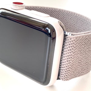 Puede incluir: Un Apple Watch negro con una banda blanca. El reloj tiene una esfera negra y un botón rojo en el lateral.