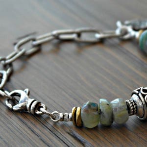 Könnte beinhalten: Ein silbernes Armband mit einer Mischung aus Kettengliedern und Edelsteinperlen. Das Armband verfügt über einen Karabinerverschluss und dekorative silberne Perlen. Die Edelsteine sind eine Mischung aus grünen und blauen Farbtönen.