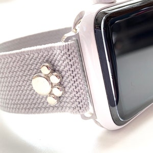 Puede incluir: Una banda de Apple Watch gris con un dije de pata de animal plateado. La banda está hecha de un material suave y elástico y es compatible con Apple Watch Series 4 y posteriores.