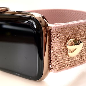 May include: A black Apple Watch with a rose gold band. The band is a stretchy, pink fabric with a gold heart-shaped accent.