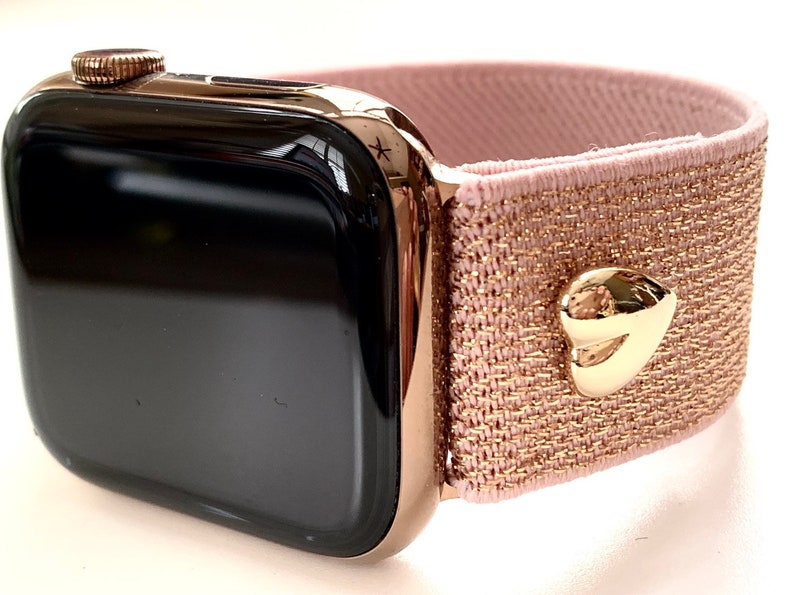May include: A black Apple Watch with a rose gold band. The band is a stretchy, pink fabric with a gold heart-shaped accent.