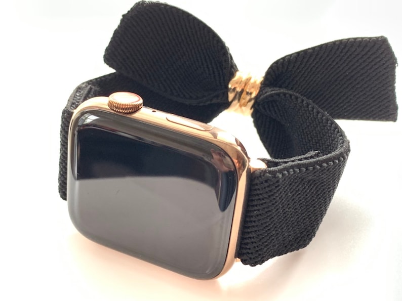 May include: A rose gold Apple Watch with a black woven band and a black fabric bow.