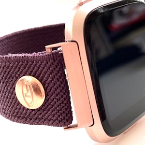May include: Rose gold smartwatch with a dark purple fabric band. The band has a rose gold button with a logo on it. The watch face is black and says "Fitbit" on the side.