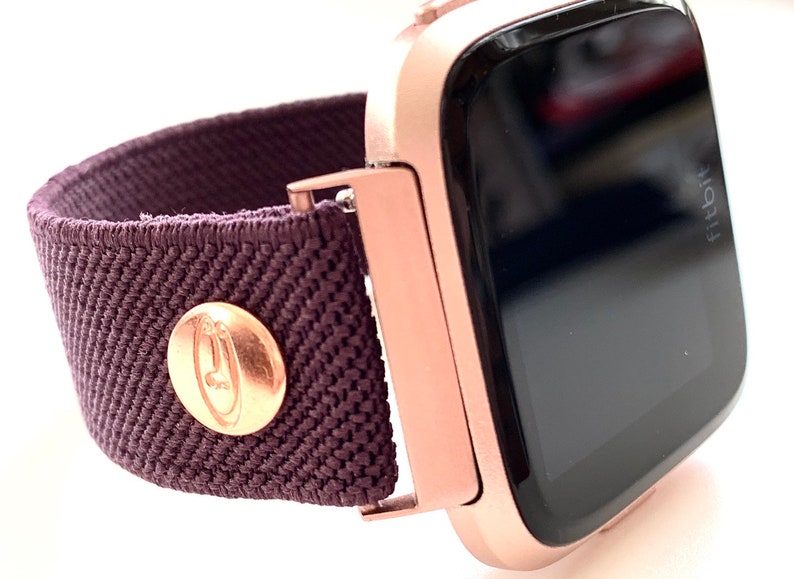 May include: Rose gold smartwatch with a dark purple fabric band. The band has a rose gold button with a logo on it. The watch face is black and says "Fitbit" on the side.