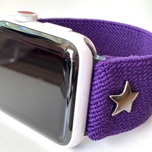May include: A white Apple Watch with a purple elastic band and a silver star charm.