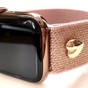 May include: A rose gold Apple Watch with a pink and gold glitter elastic band. The band has a gold heart-shaped buckle.