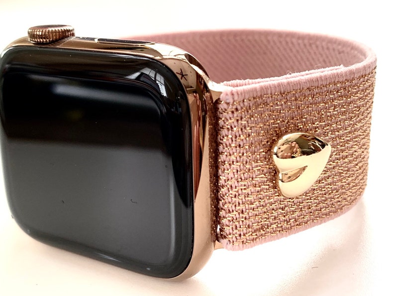 May include: A rose gold Apple Watch with a pink and gold glitter elastic band. The band has a gold heart-shaped buckle.