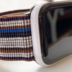 Puede incluir: Un Apple Watch blanco con una pantalla negra y una banda tejida a rayas de colores. La banda tiene rayas marrones, blancas, azules y moradas.