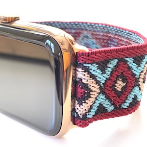 Op de afbeelding: Een Apple Watch in rosé goud met een kleurrijk geweven bandje. Het bandje heeft een geometrisch patroon in tinten rood, blauw, beige en zwart.