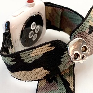 May include: A white Apple Watch with a camouflage patterned elastic band and a silver skull charm.