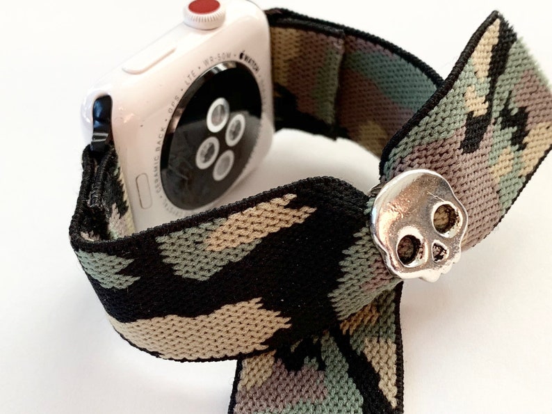 May include: A white Apple Watch with a camouflage patterned elastic band and a silver skull charm.