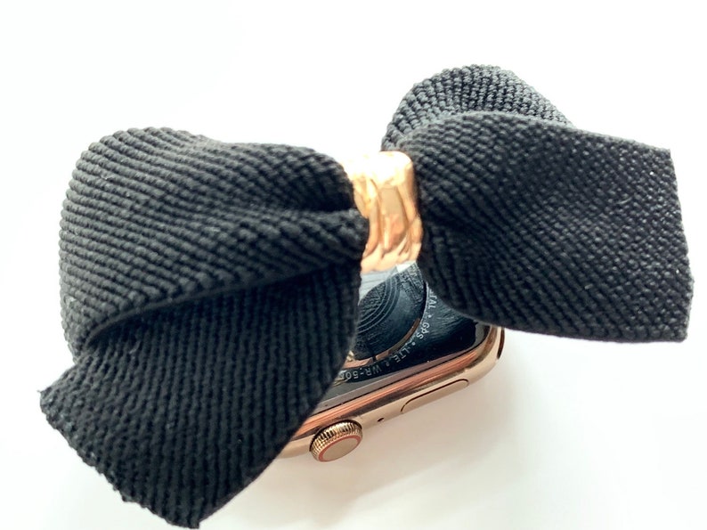 May include: A black fabric bow with a gold metal band is attached to a gold Apple Watch. The watch face is black and the text "WATER RESISTANT 50M" is visible on the watch band.
