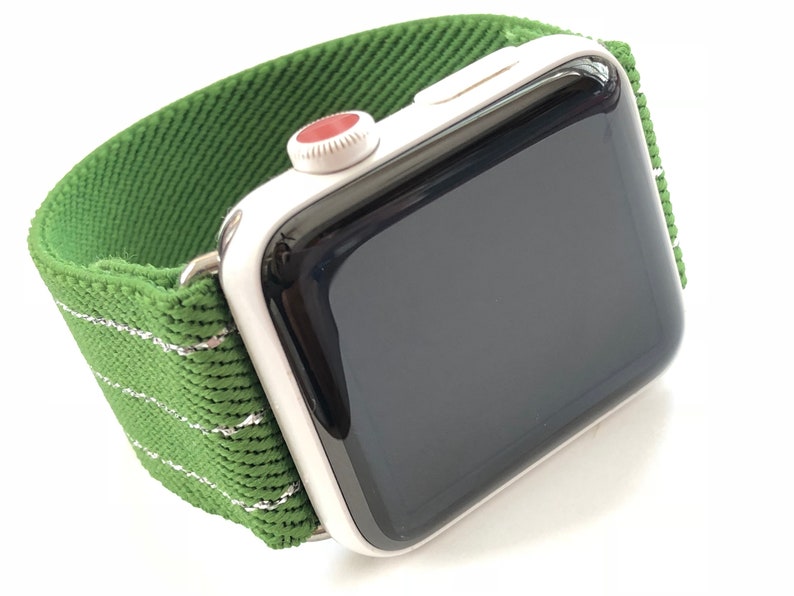 Puede incluir: Un Apple Watch plateado con una banda deportiva de rayas verdes y blancas. La esfera del reloj es negra y la corona digital es roja.