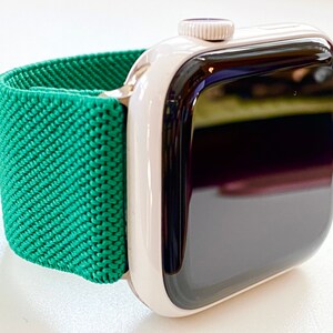 K&ouml;nnte beinhalten: Eine wei&szlig;e Apple Watch mit einem gr&uuml;nen Sportarmband. Das Zifferblatt der Uhr ist schwarz und das Armband besteht aus einem weichen, dehnbaren Material.