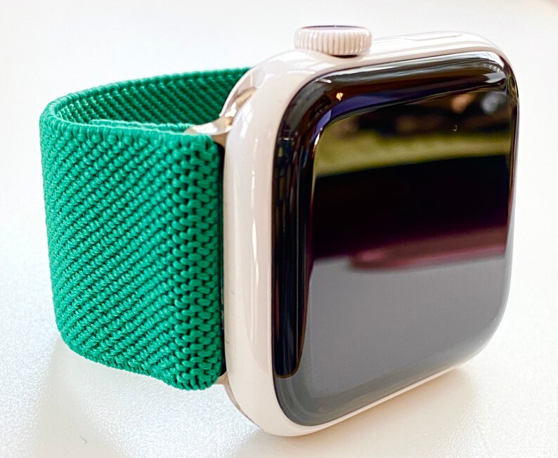 K&ouml;nnte beinhalten: Eine wei&szlig;e Apple Watch mit einem gr&uuml;nen Sportarmband. Das Zifferblatt der Uhr ist schwarz und das Armband besteht aus einem weichen, dehnbaren Material.