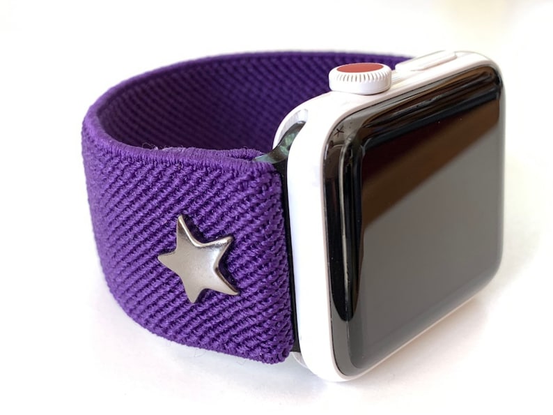 May include: A purple elastic Apple Watch band with a silver star charm. The watch band is compatible with Apple Watch Series 4 and later.