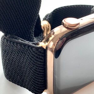 May include: A rose gold Apple Watch with a black woven band. The watch face is black and the band is made of a soft, stretchy material.