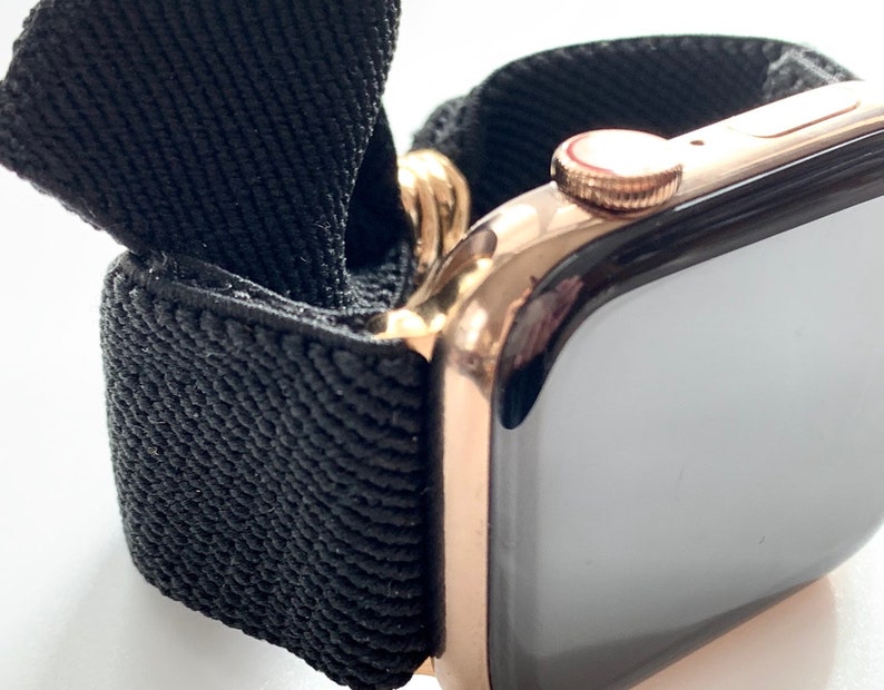 May include: A rose gold Apple Watch with a black woven band. The watch face is black and the band is made of a soft, stretchy material.