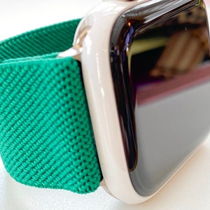 K&ouml;nnte beinhalten: Eine wei&szlig;e Apple Watch mit einem gr&uuml;nen Sportarmband. Das Zifferblatt der Uhr ist schwarz und reflektierend.