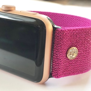 May include: A rose gold Apple Watch with a pink sparkly sport band. The band has a small rhinestone embellishment.